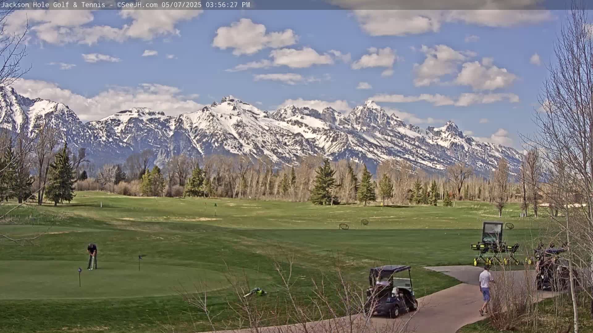 A golf course with golfers and golf carts is situated in front of a snow-capped mountain range under a partly cloudy sky.