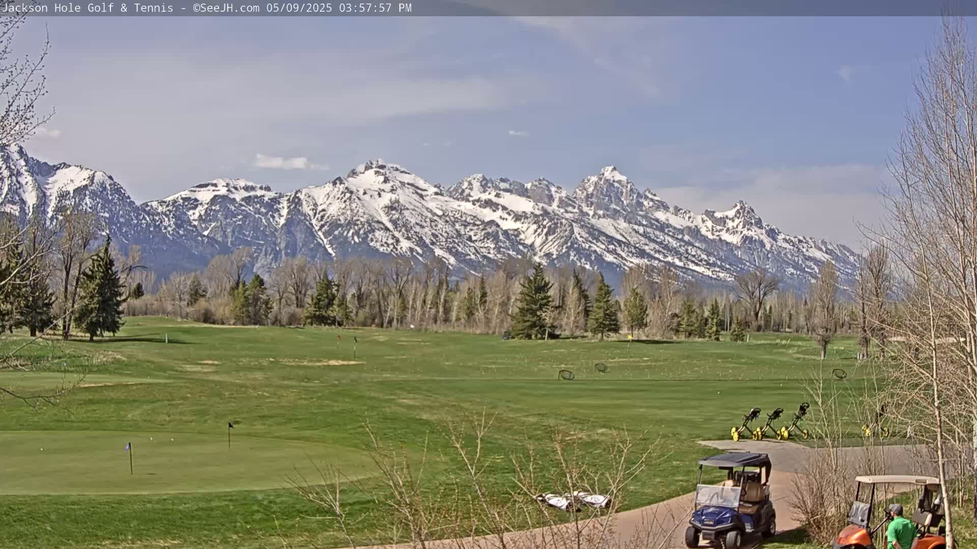 A golf course with snow-capped mountains in the background under a mostly clear sky.