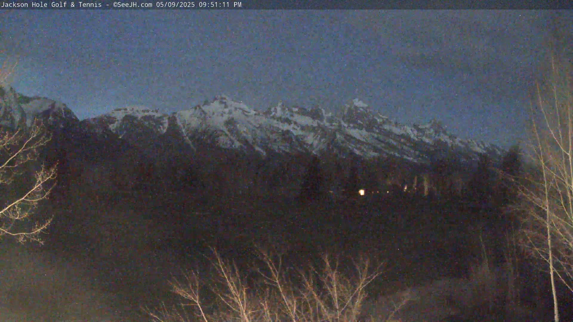 Snow-capped mountains are visible in the distance under a twilight sky, with bare trees and a few distant lights in the foreground.