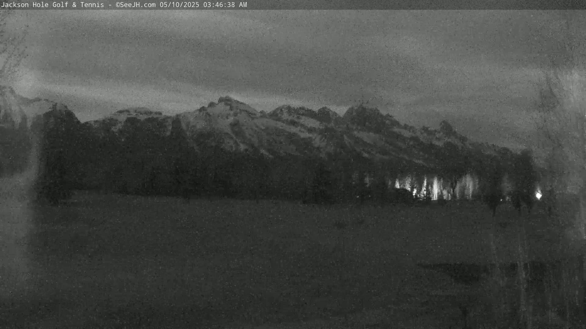 A nighttime, low-light image shows snow-capped mountains in the distance, with a line of trees and a few lights visible in the foreground.