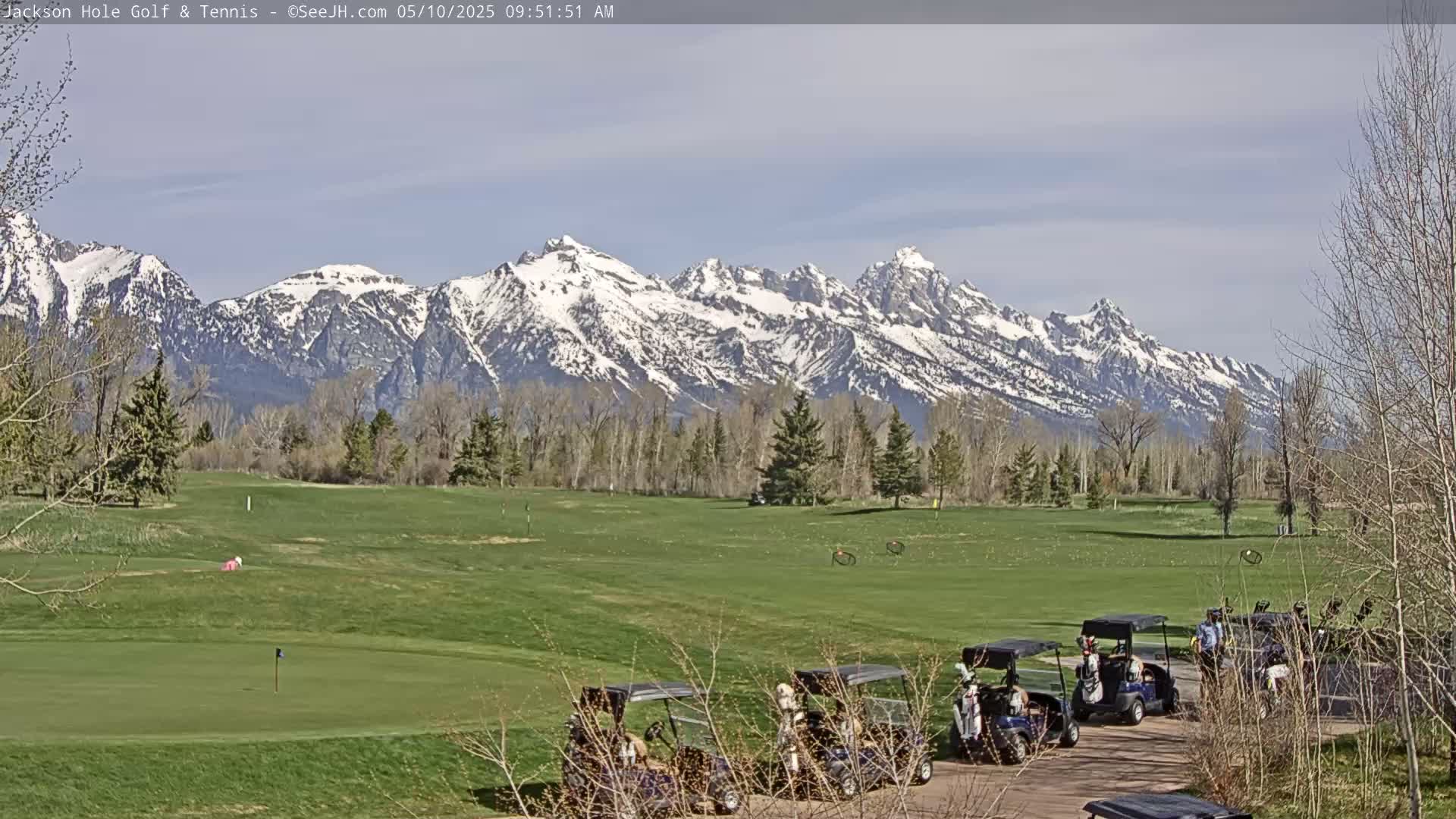 A golf course with several golf carts parked near the clubhouse, situated in front of a snow-capped mountain range under a mostly clear sky.