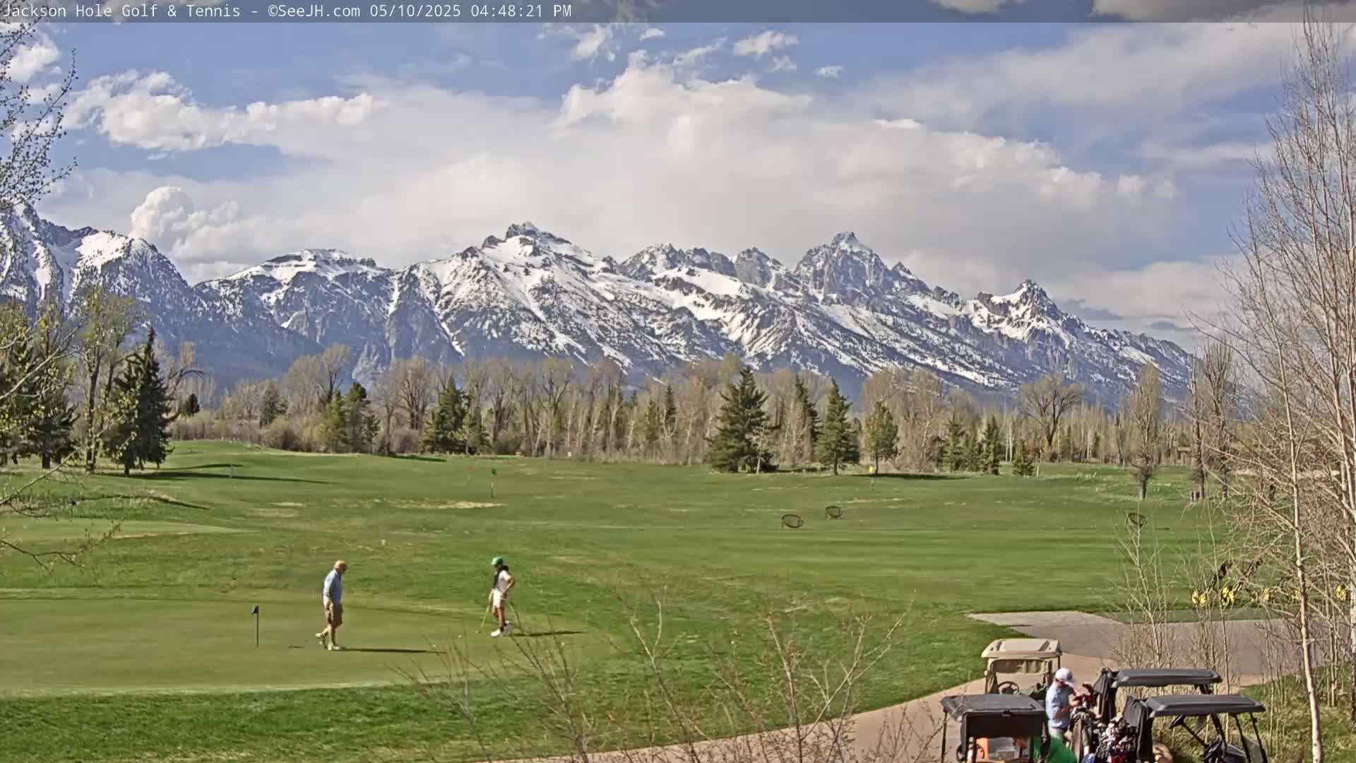 A partly cloudy day shows a golf course with several people golfing, set against a backdrop of snow-capped mountains.