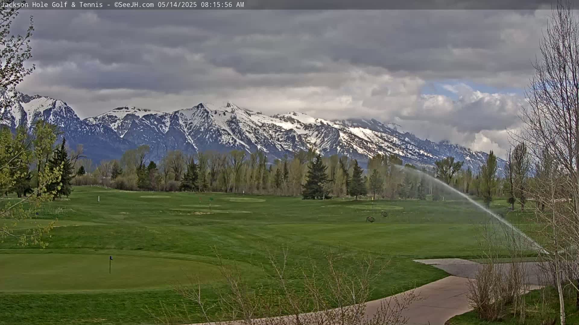 A golf course with a sprinkler system running is situated in front of a snow-capped mountain range under a cloudy sky.