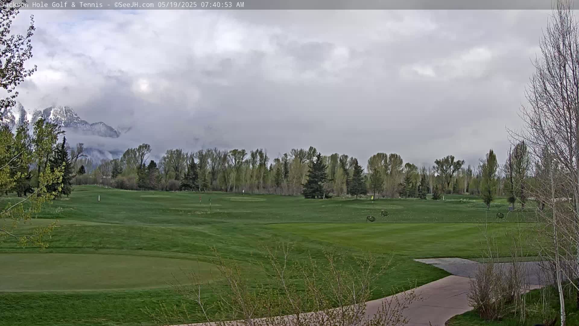 A partly cloudy day reveals a green golf course with trees and snow-capped mountains in the background.