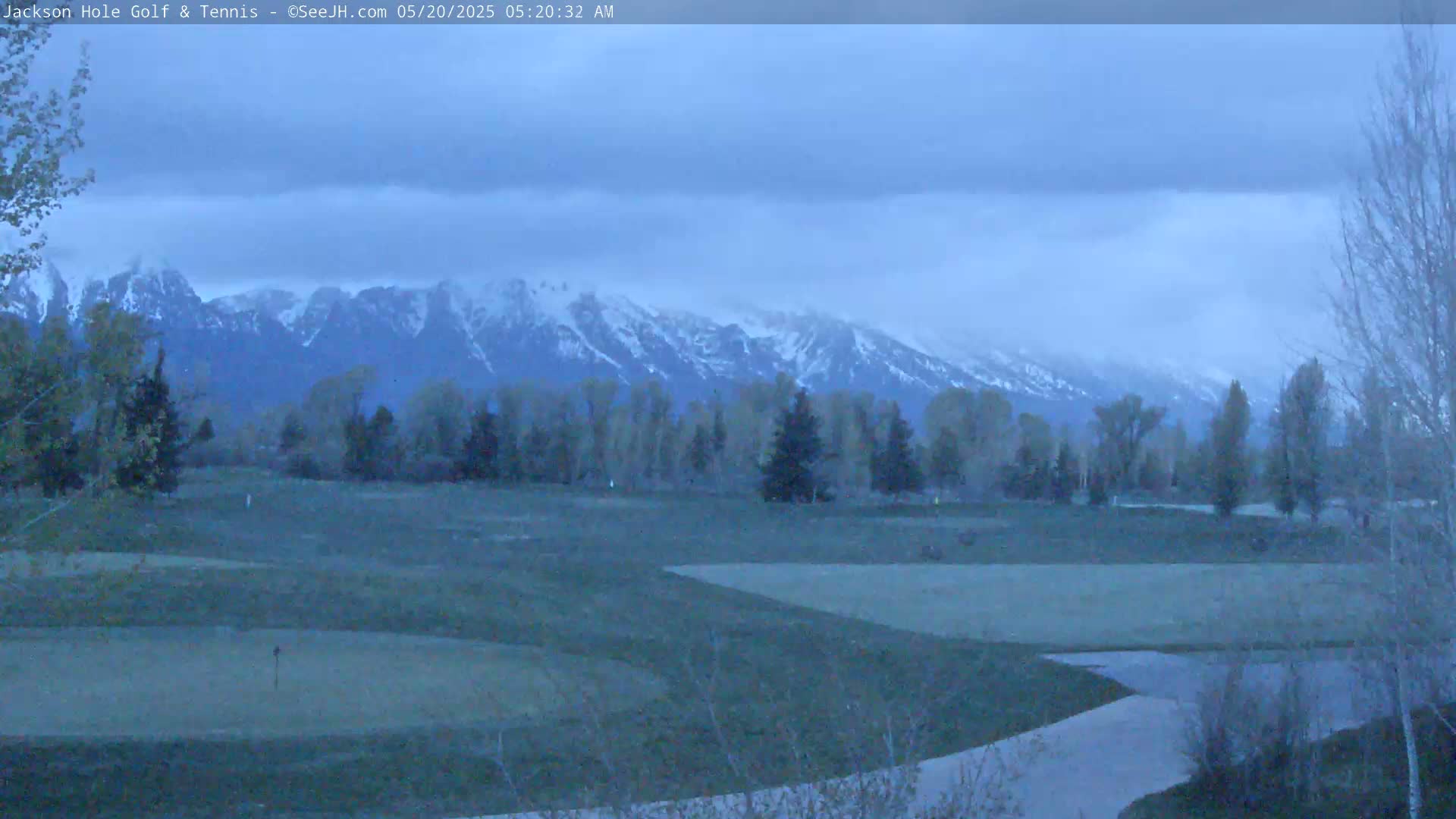 A golf course sits in the foreground, with snow-capped mountains in the background under a cloudy, dusky sky.