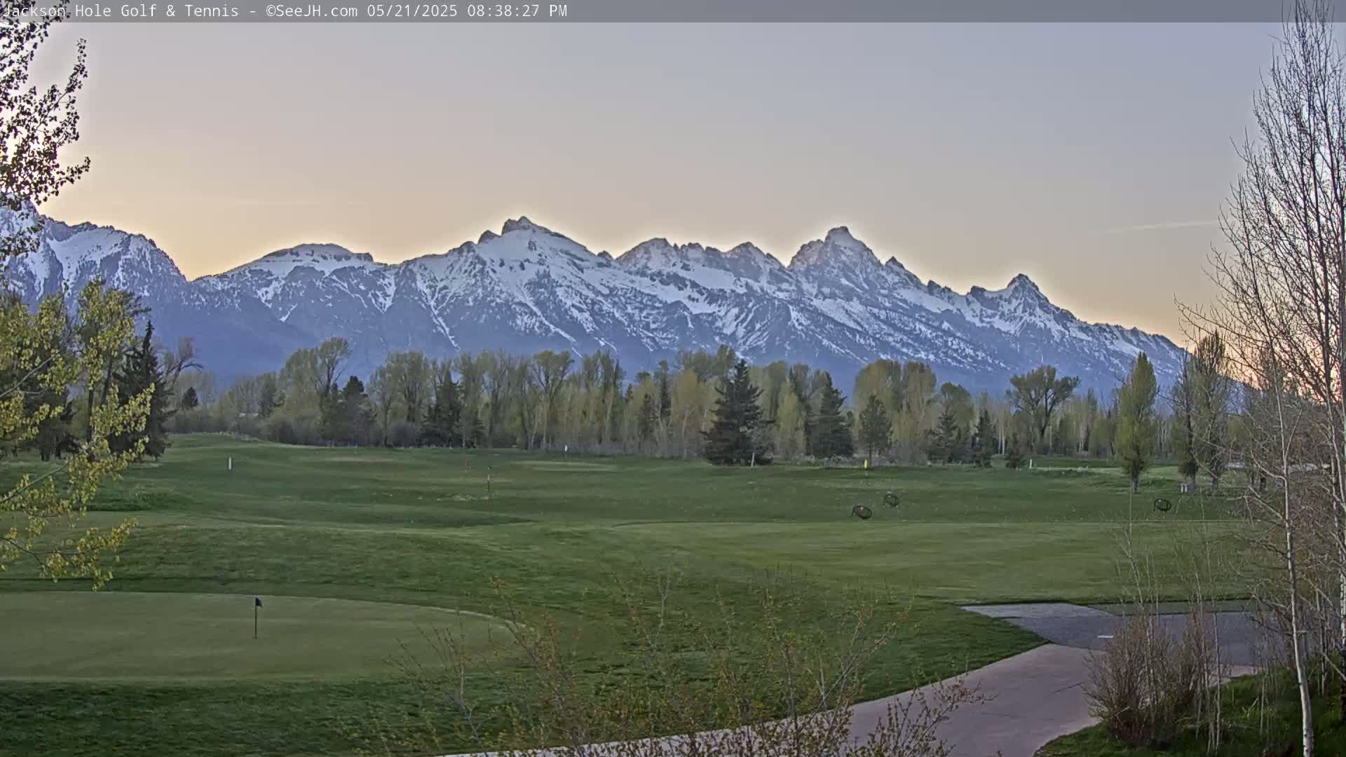 A golf course with a putting green and trees in the foreground, overlooks a snow-capped mountain range under a twilight sky.