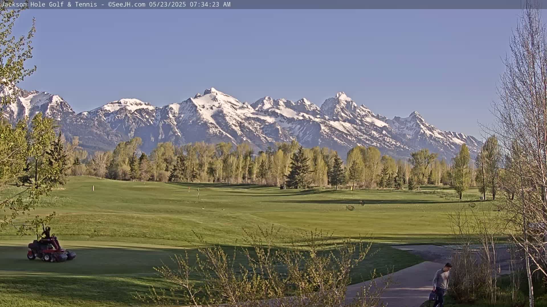 A golf course with a person walking and a groundskeeping vehicle is visible in the foreground, with a snow-capped mountain range under a clear blue sky in the background.