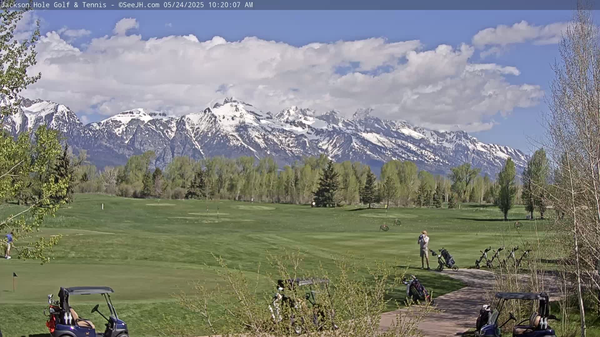 A partly cloudy day on a golf course shows snow-capped mountains in the background, with several golf carts parked near a path.