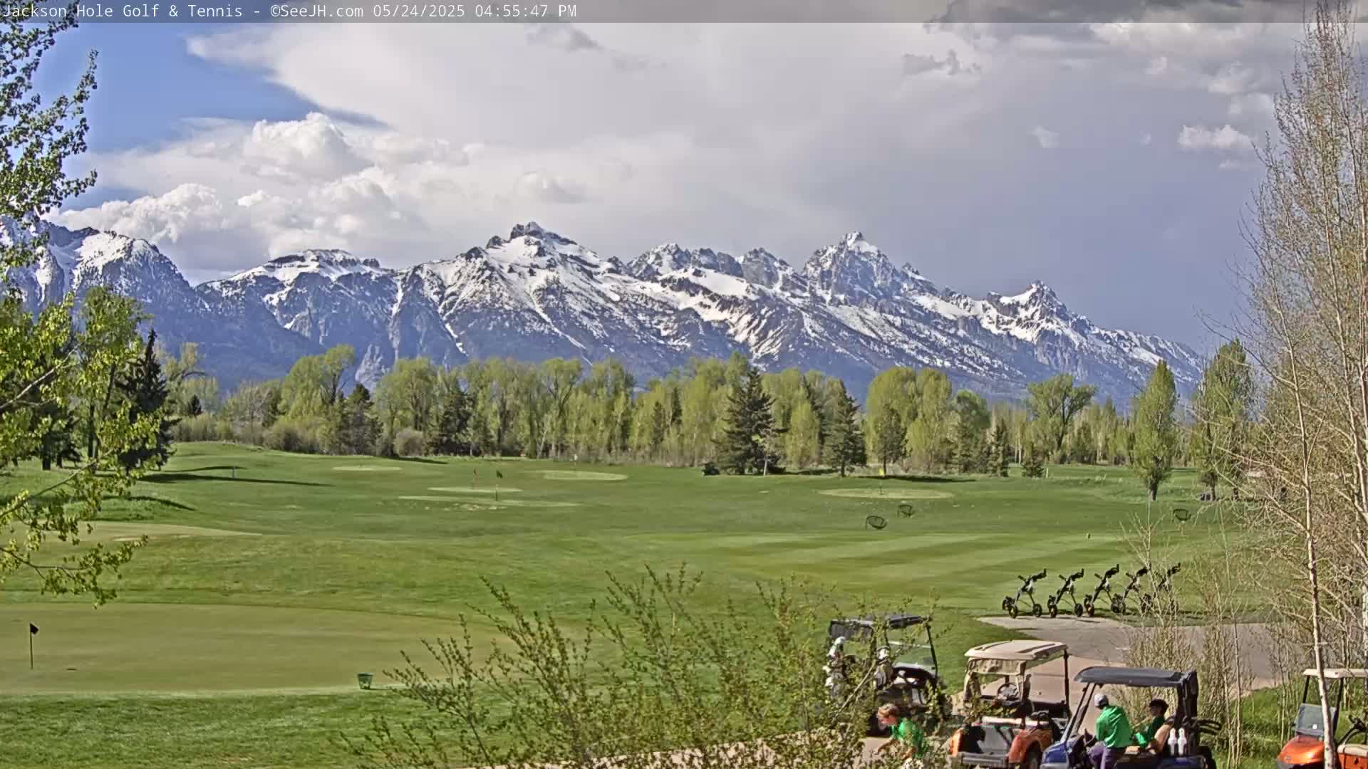A partly cloudy day shows a snow-capped mountain range in the background of a green golf course with several golf carts and people near the bottom.