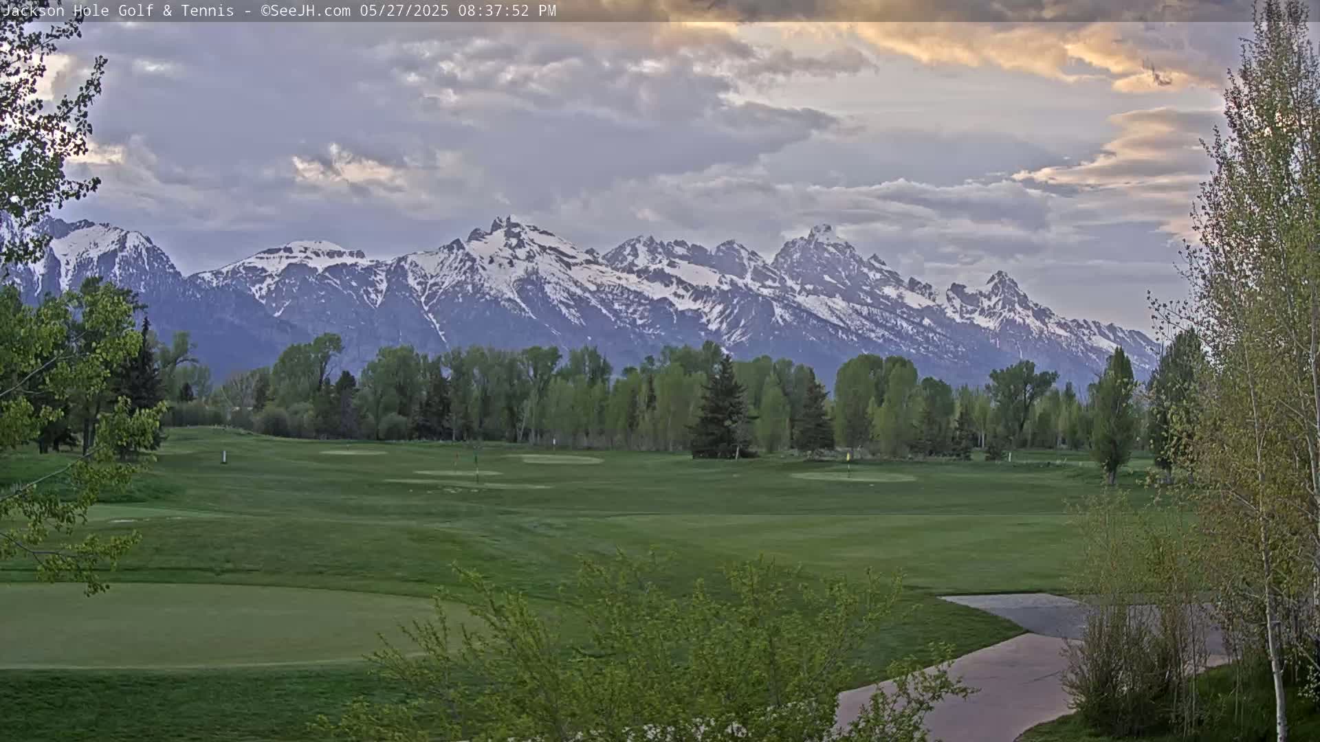 A golf course with green fairways and trees stretches out before a mountain range with snow-capped peaks under a partly cloudy sky.