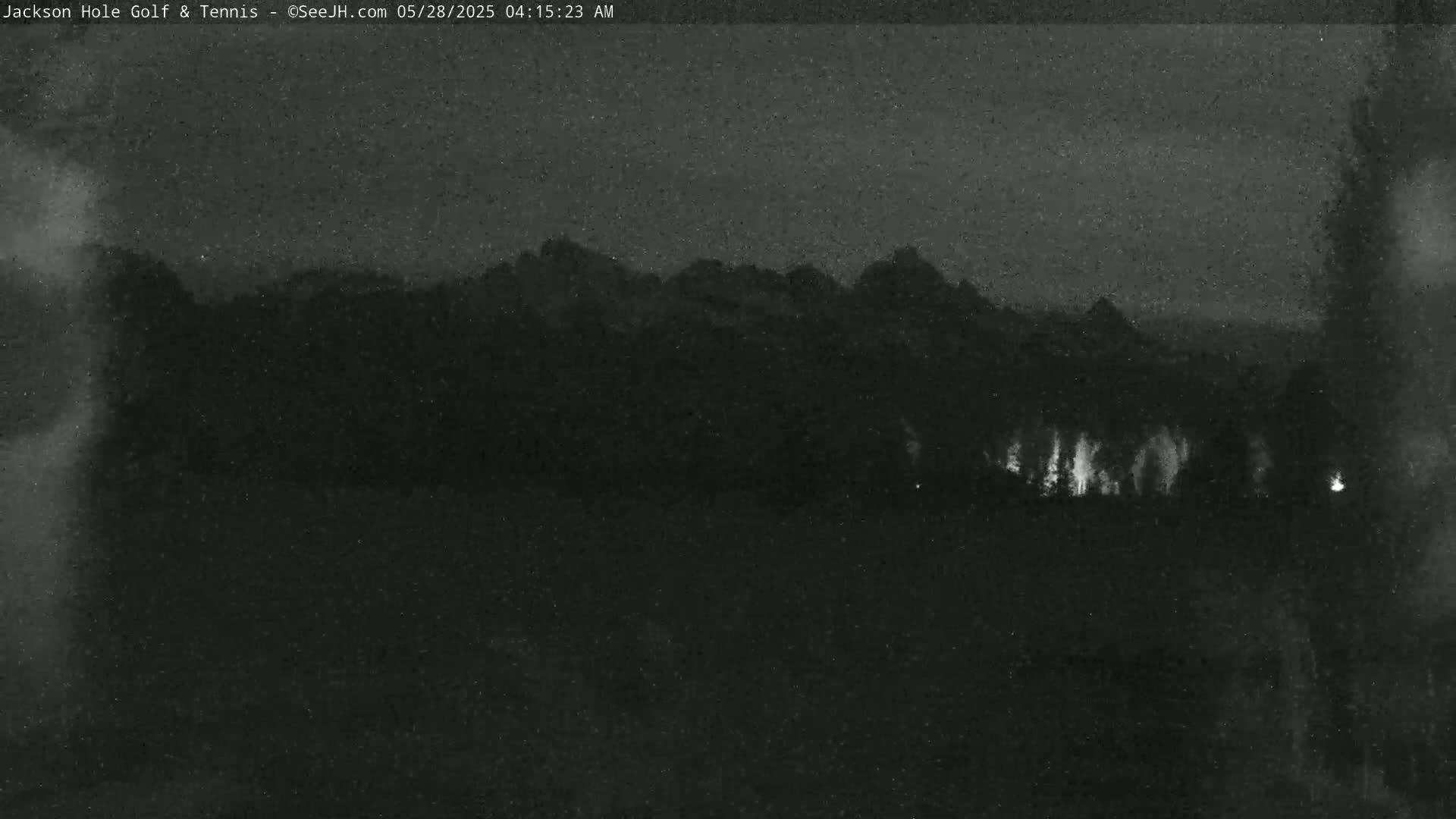 A nighttime, low-light image shows a dark landscape with a mountain range in the distance and faintly lit trees in the foreground.
