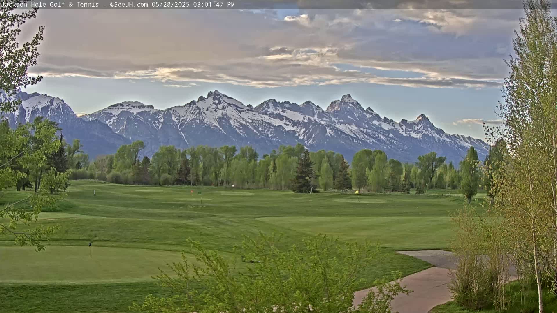 A partly cloudy sky overlooks a green golf course with trees and a snow-capped mountain range in the background.