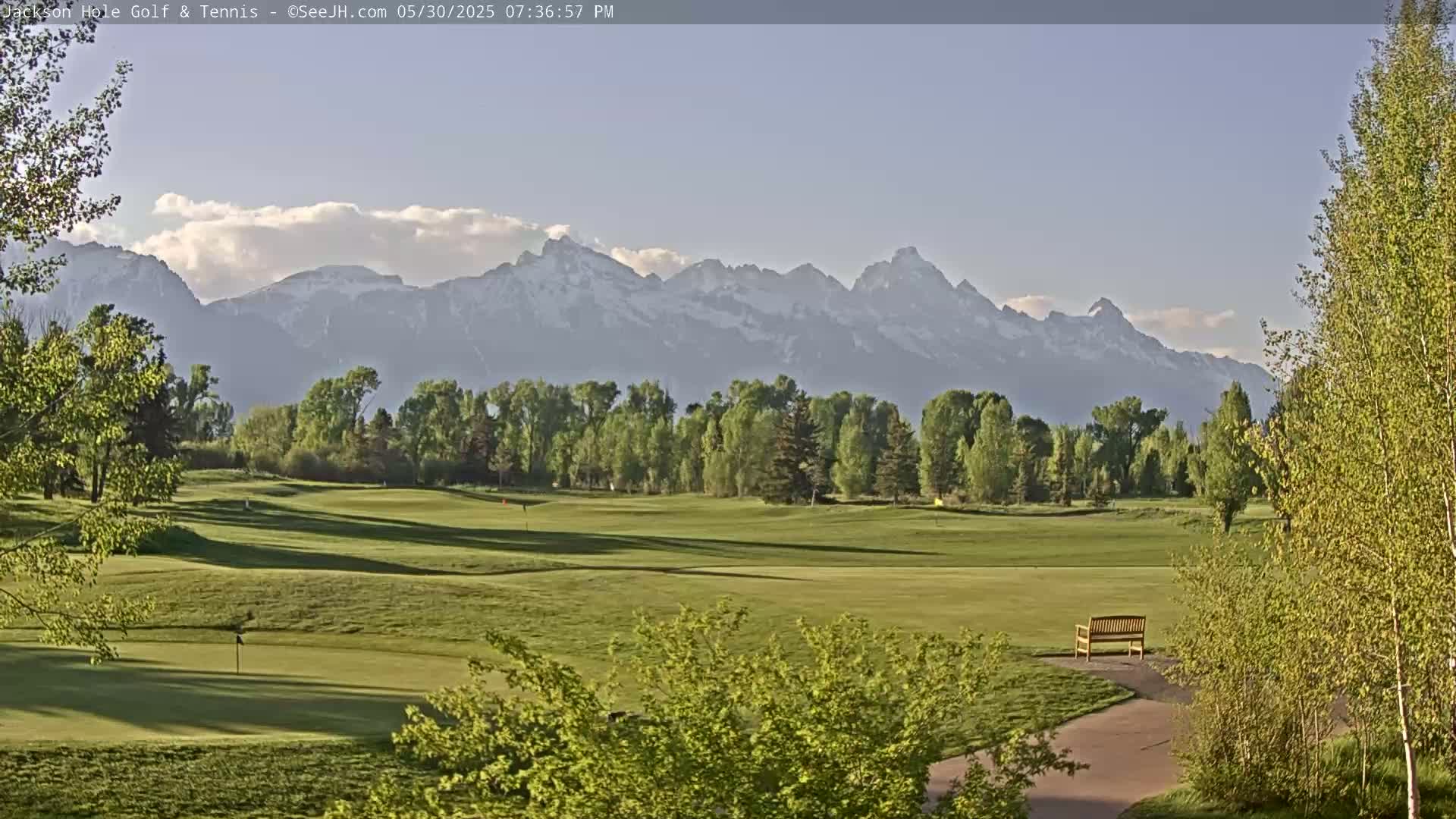 A golf course with a bench sits in the foreground, framed by green trees and a background of snow-capped mountains under a mostly clear sky.