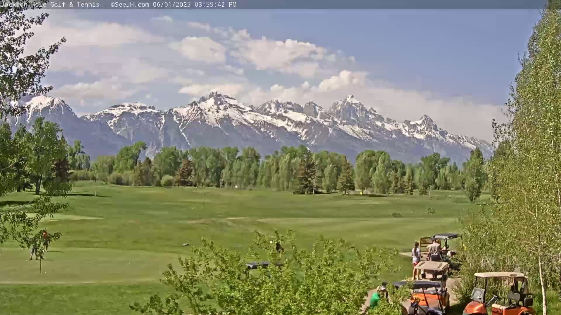 A golf course with several people and golf carts is situated in front of a snow-capped mountain range under a partly cloudy sky.