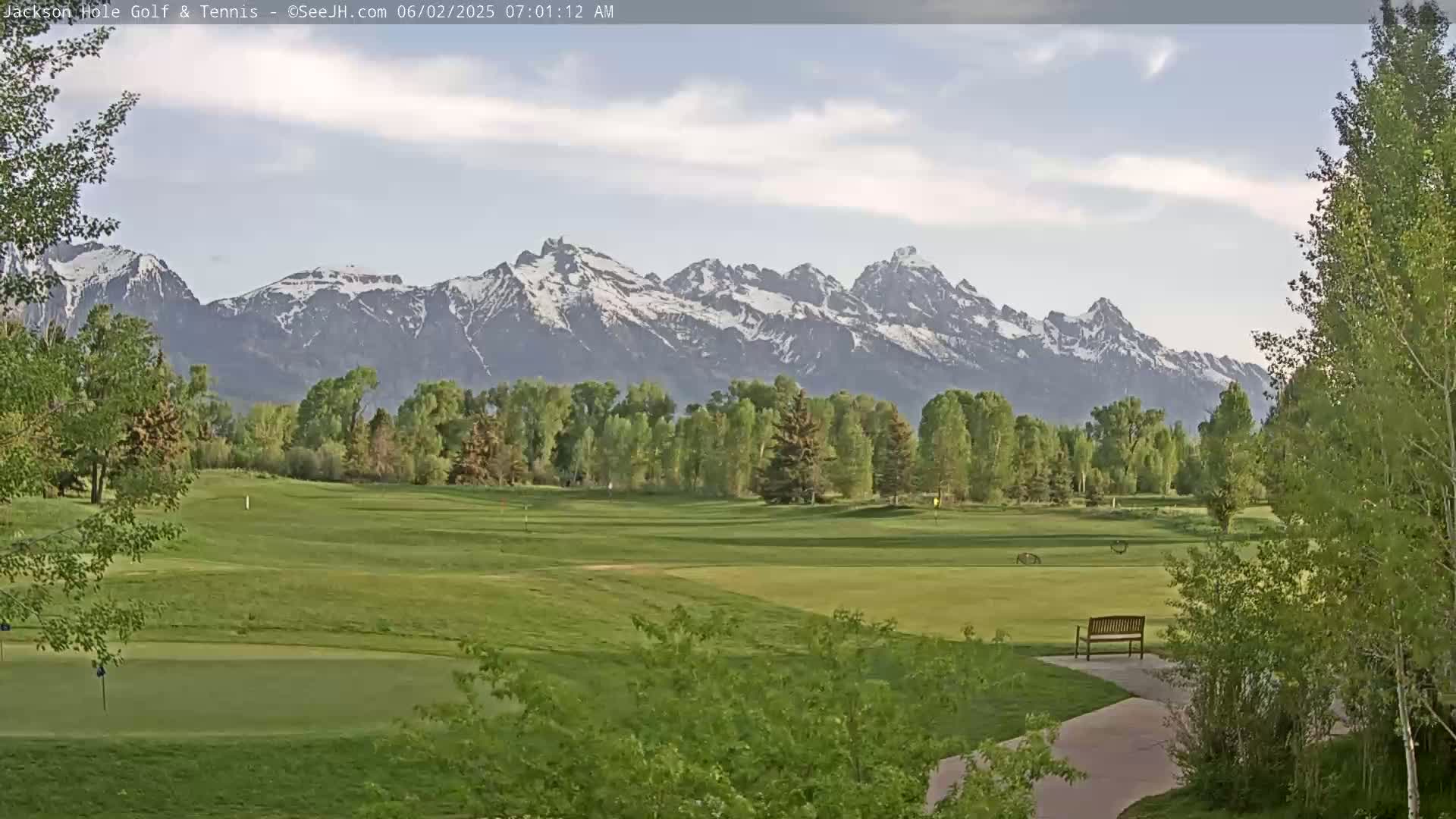 A partly cloudy morning reveals a snow-capped mountain range in the distance behind a lush green golf course with a lone bench.