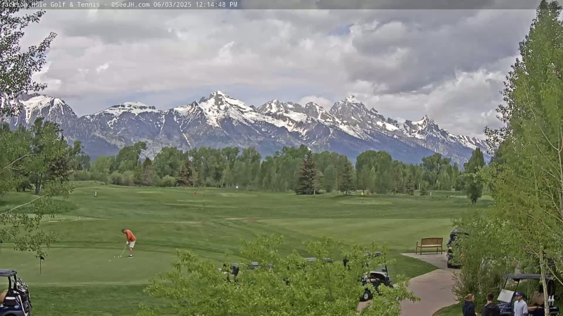 A golfer tees off on a green golf course with snow-capped mountains in the background under a partly cloudy sky.