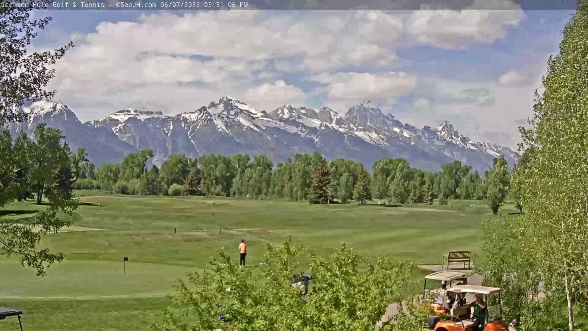 A partly cloudy day shows a golf course with a person putting, golf carts, and a mountain range in the background.