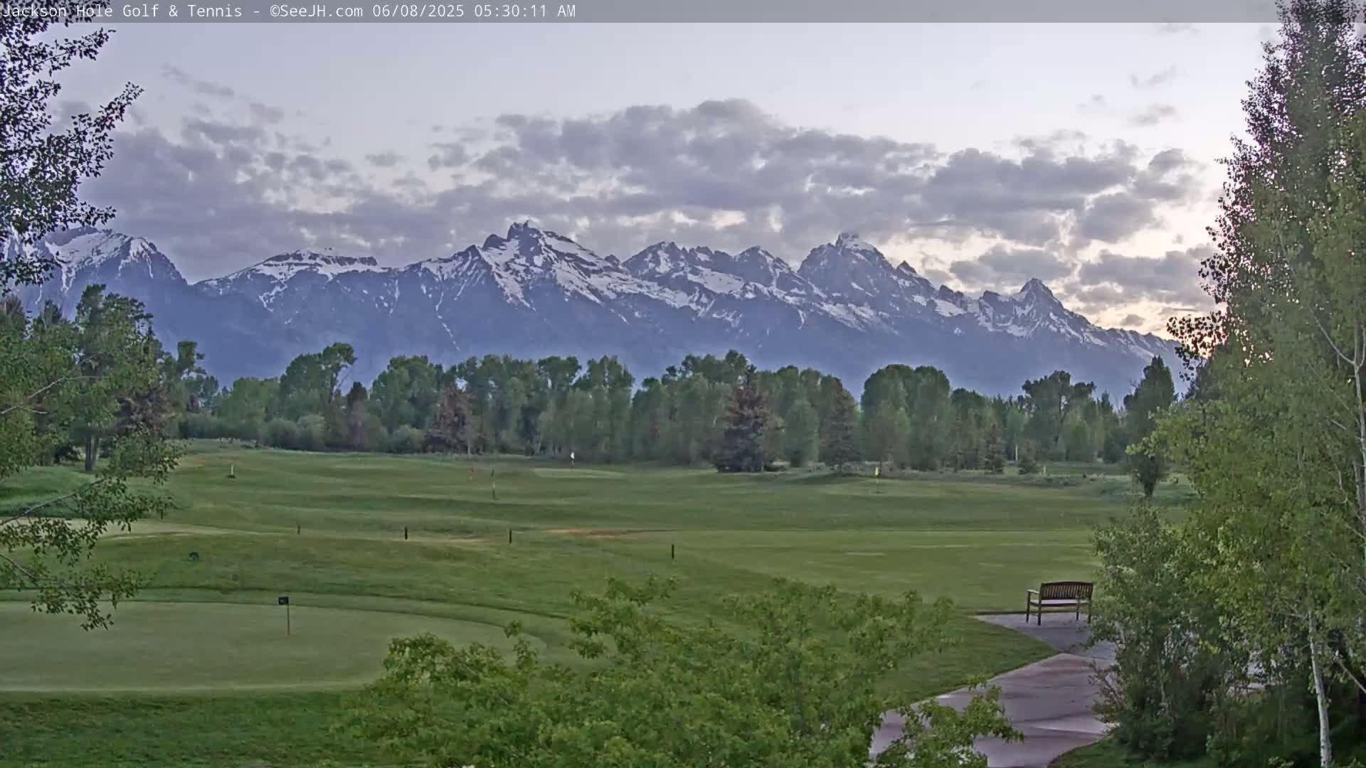 A golf course with a bench sits before a backdrop of snow-capped mountains under a partly cloudy sky.