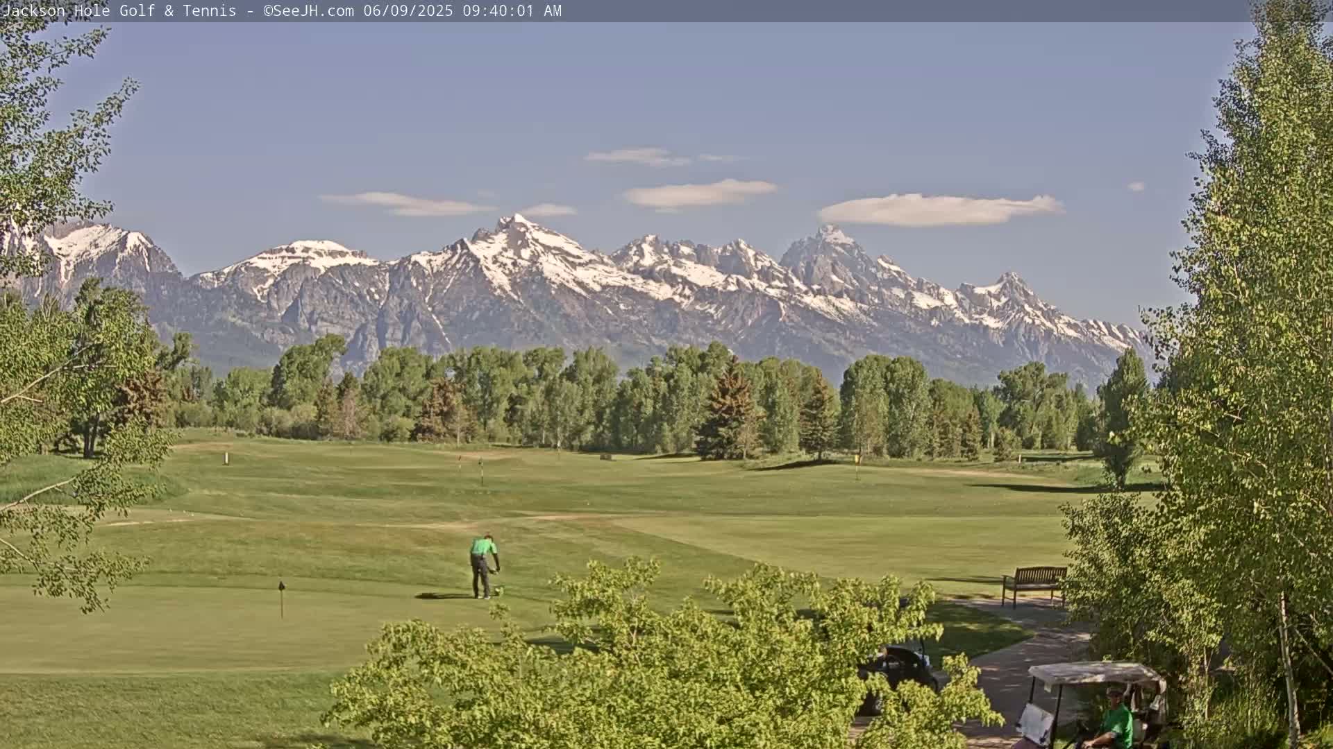 A golfer is putting on a green golf course with snow-capped mountains in the background under a mostly clear sky.