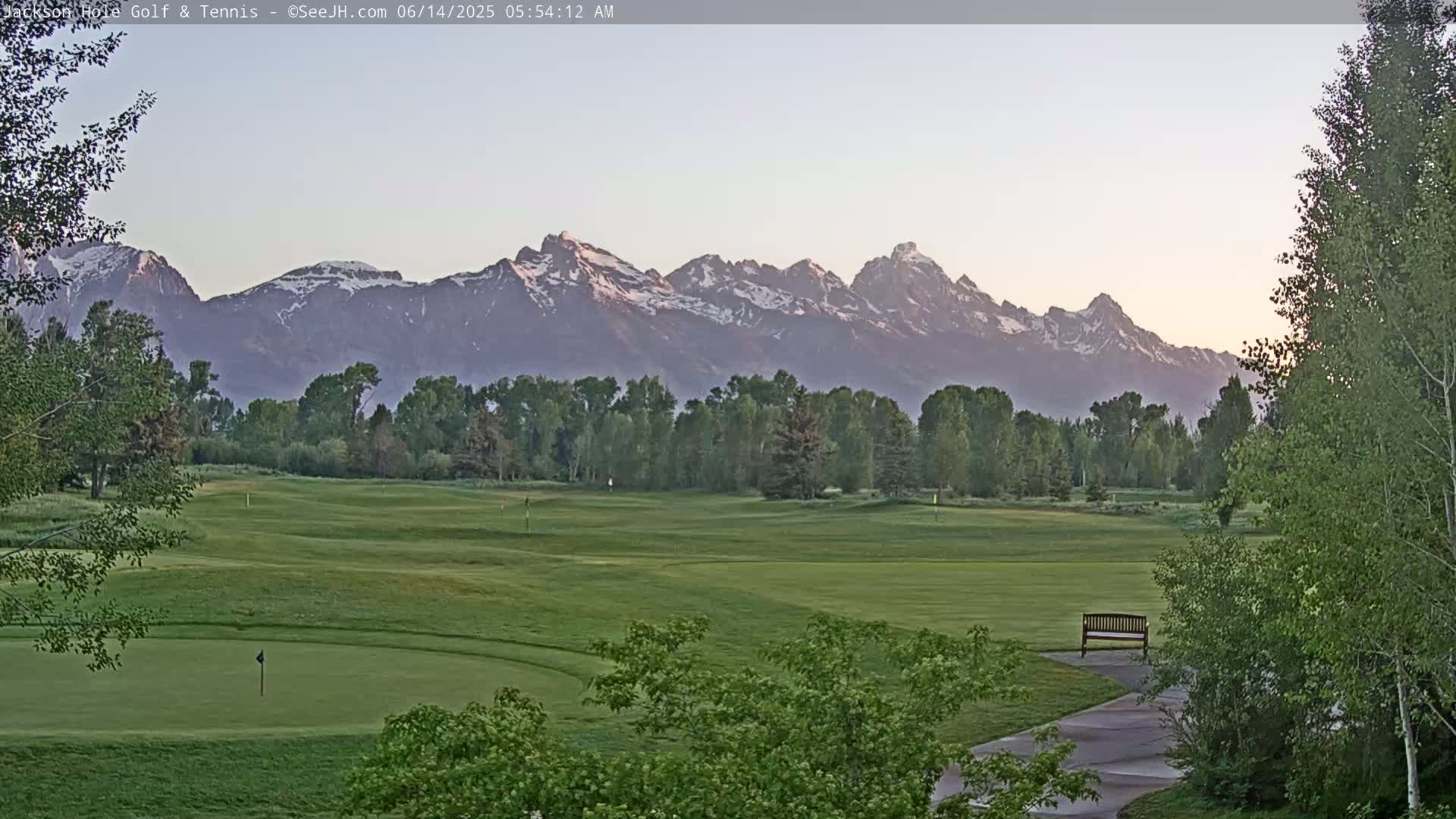 A golf course with a putting green, trees, and a bench overlooks a mountain range with snow-capped peaks under a dawn sky.