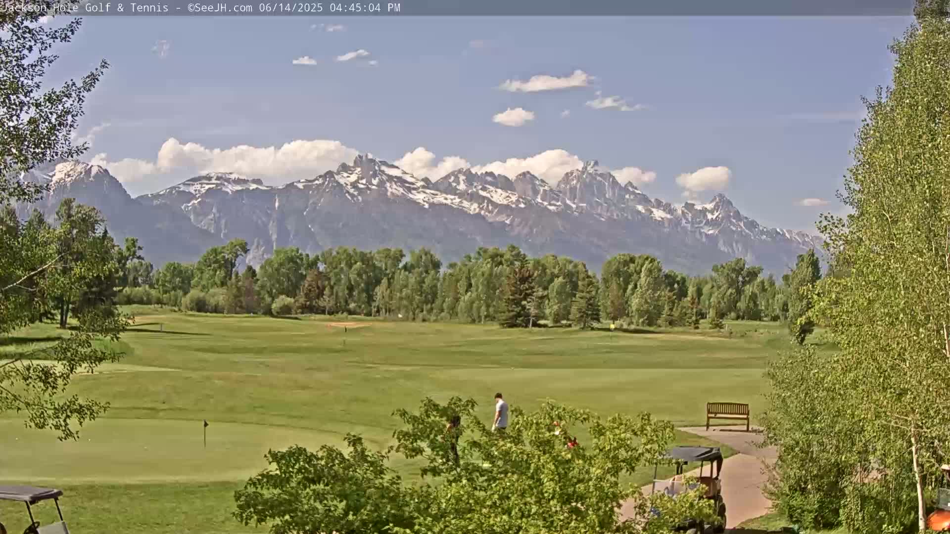 A golf course with several golf carts is situated in front of a snow-capped mountain range on a partly cloudy day.