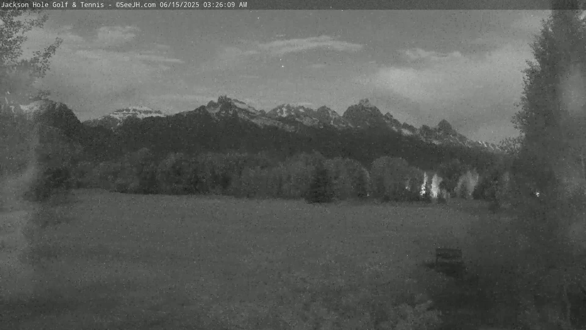 A grayscale image shows a flat field at night with trees and snow-capped mountains in the background under a mostly cloudy sky.