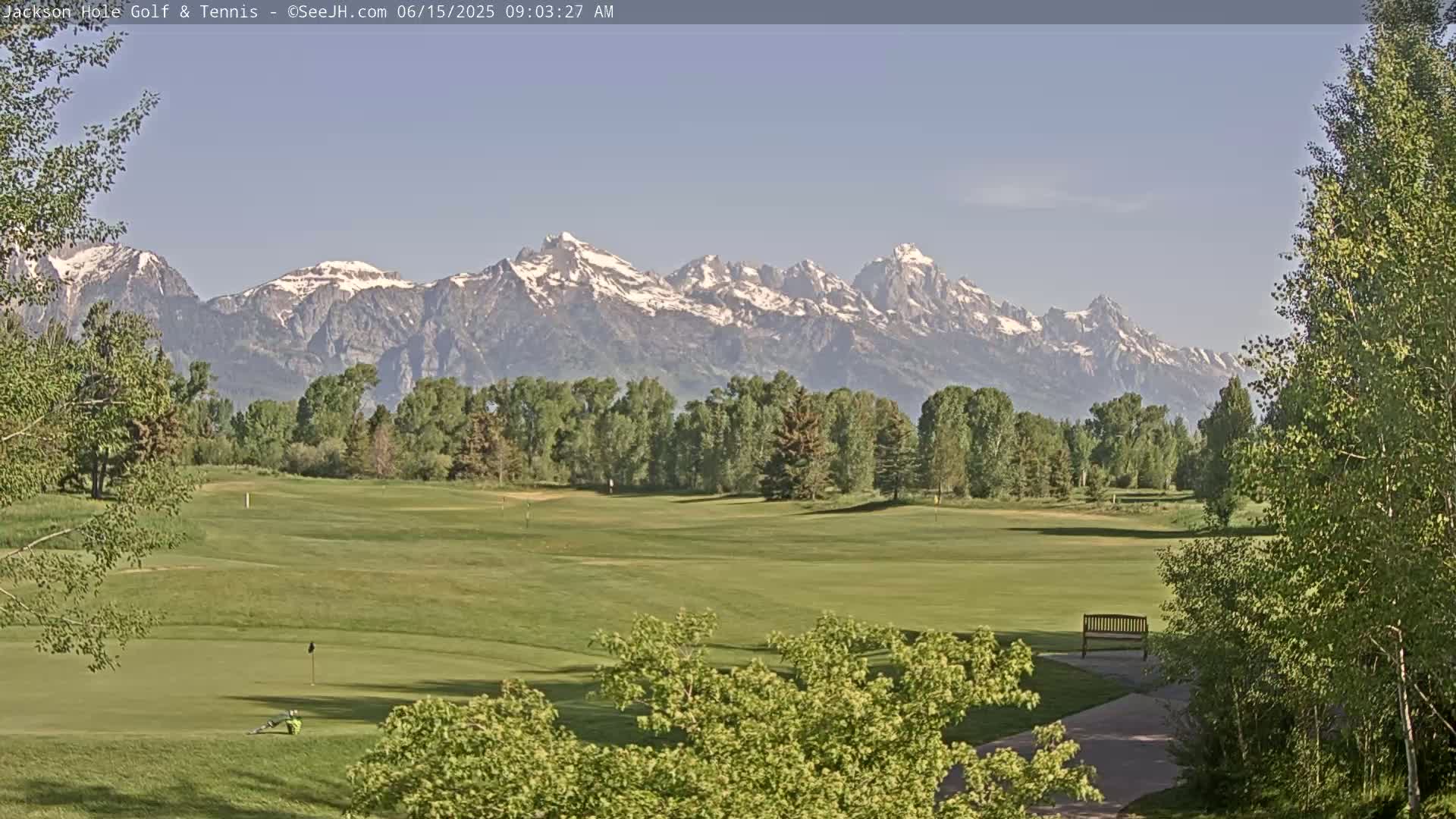 A golf course with green fairways and trees stretches towards a snow-capped mountain range under a clear sky.
