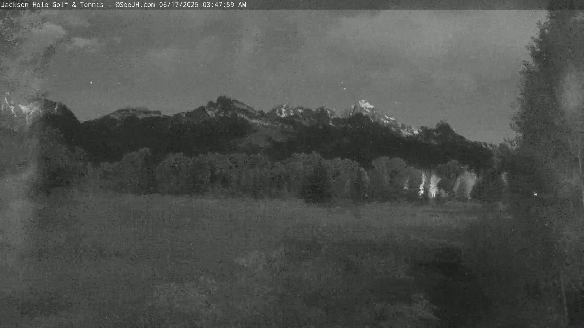 A nighttime, low-light image shows a mountainous landscape with trees in the foreground under a cloudy sky.