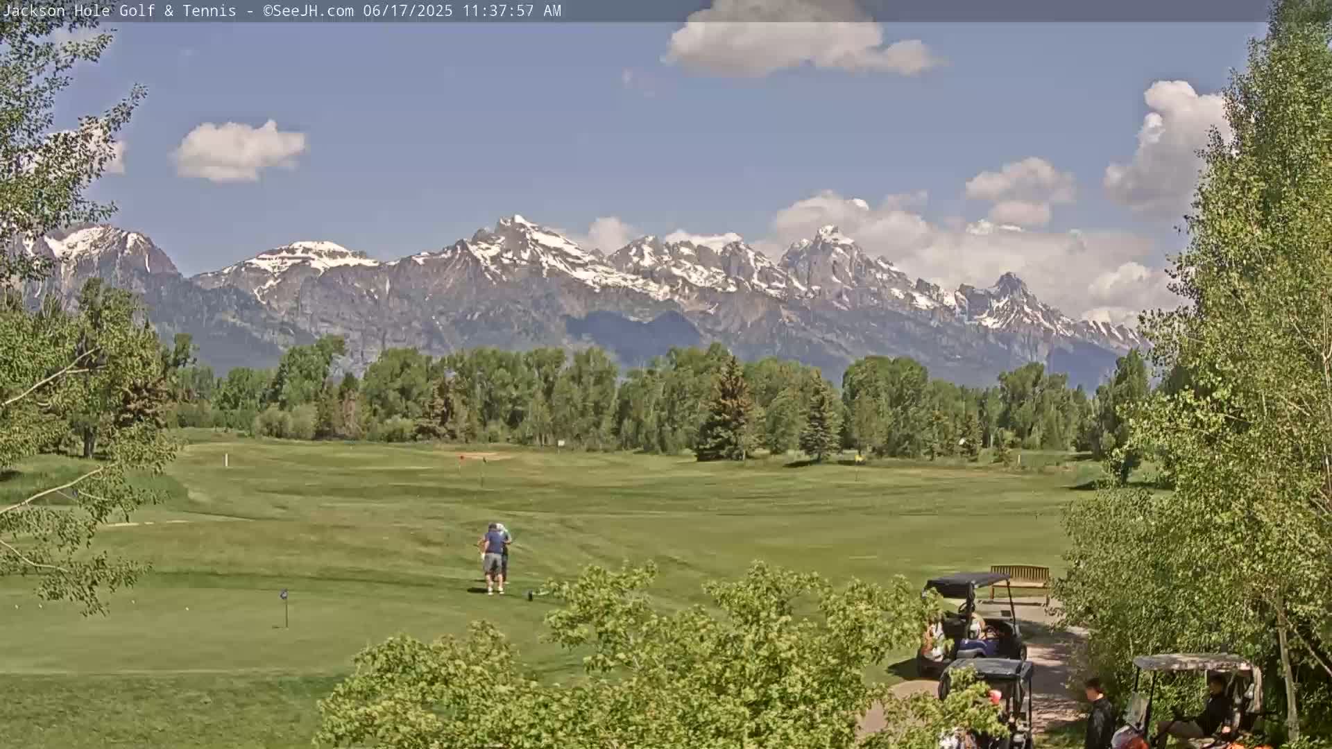 A golf course with two golfers on the fairway, bordered by green trees and overlooking a snow-capped mountain range under a partly cloudy sky.