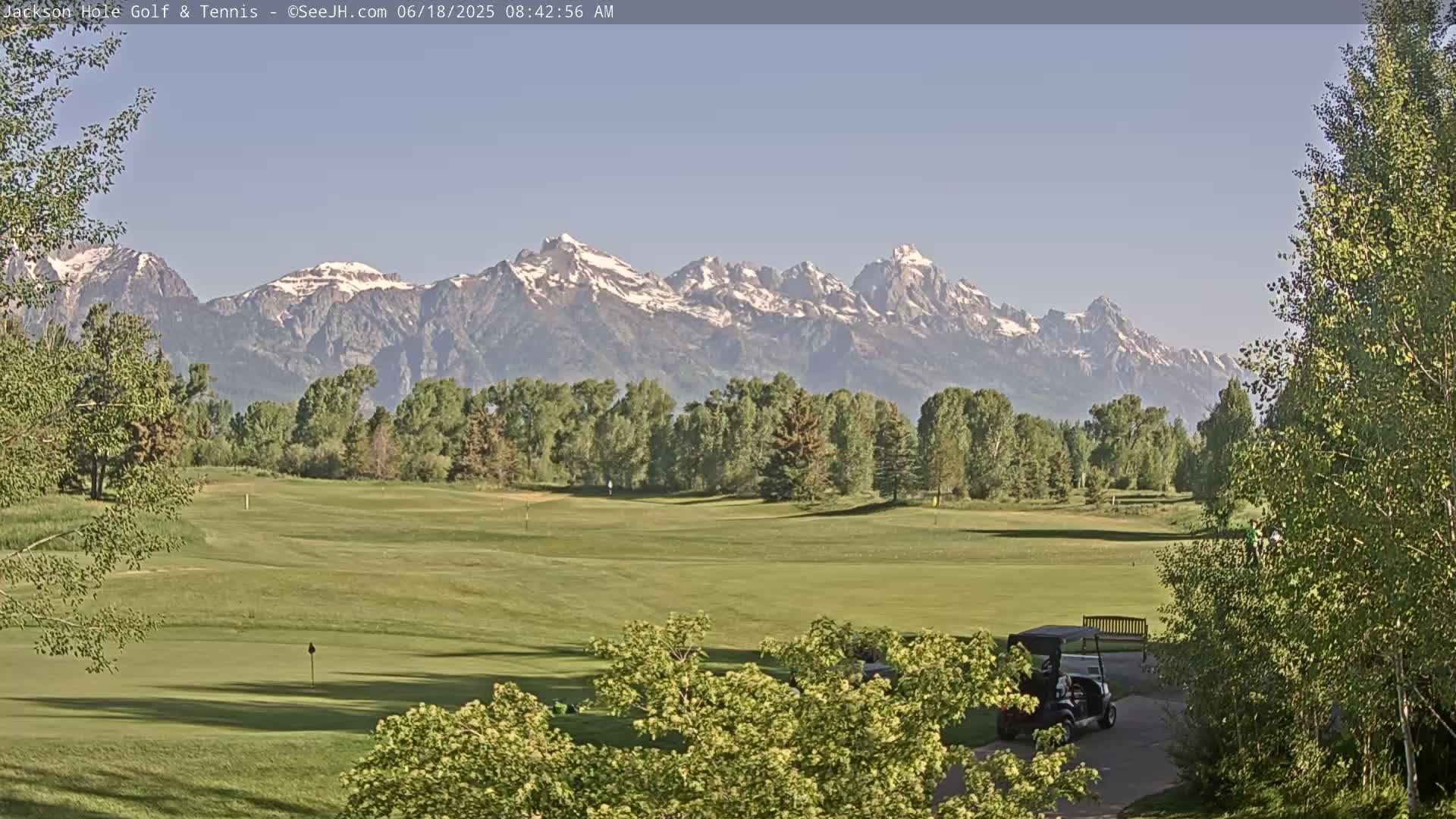 A golf course with green fairways and trees is situated in the foreground, with snow-capped mountains visible in the background under a clear sky.