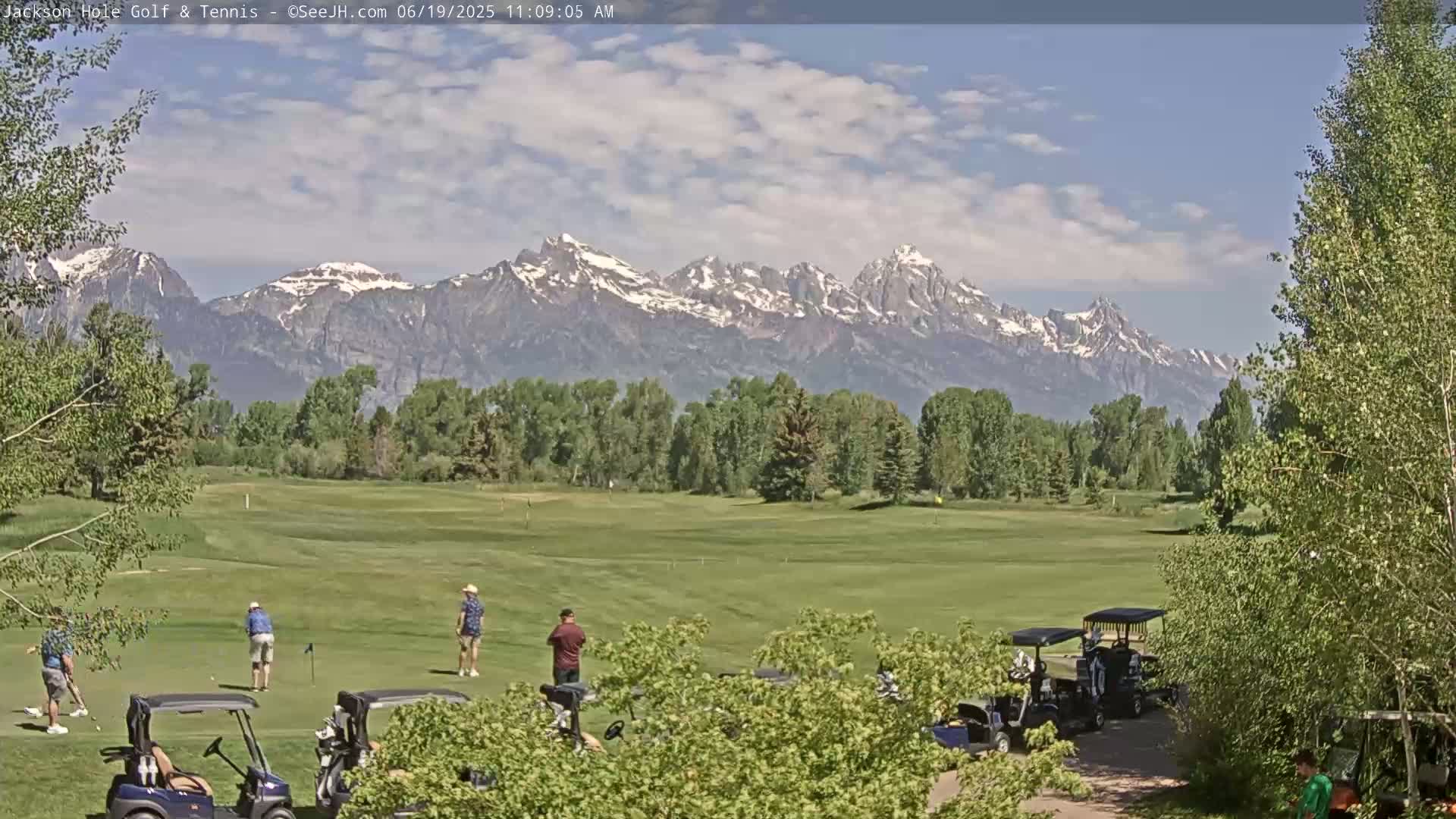 A partly sunny day on a golf course with snow-capped mountains in the background, golfers playing on the green, and golf carts parked nearby.