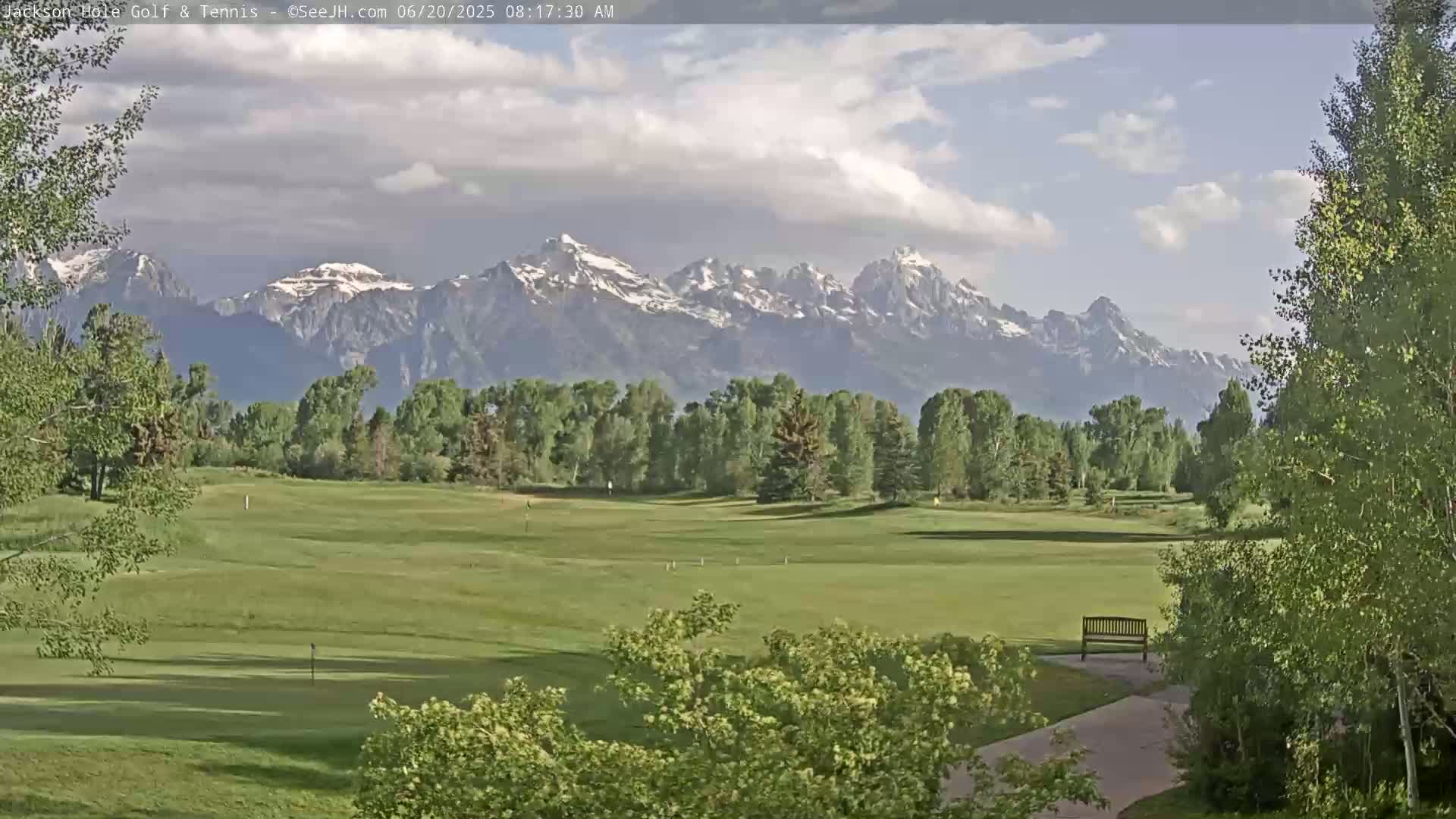 A partly cloudy sky overlooks a green golf course with trees and a bench, all situated in front of a snow-capped mountain range.