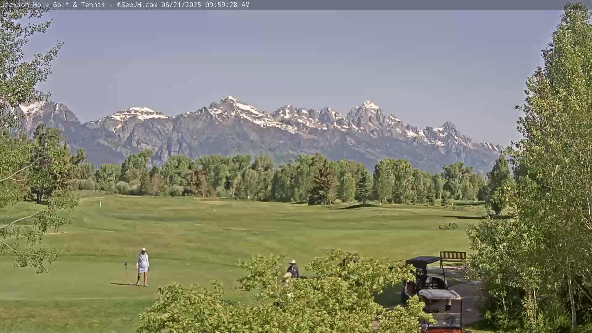 A golf course with a few people playing golf stretches out before a backdrop of snow-capped mountains under a clear sky.