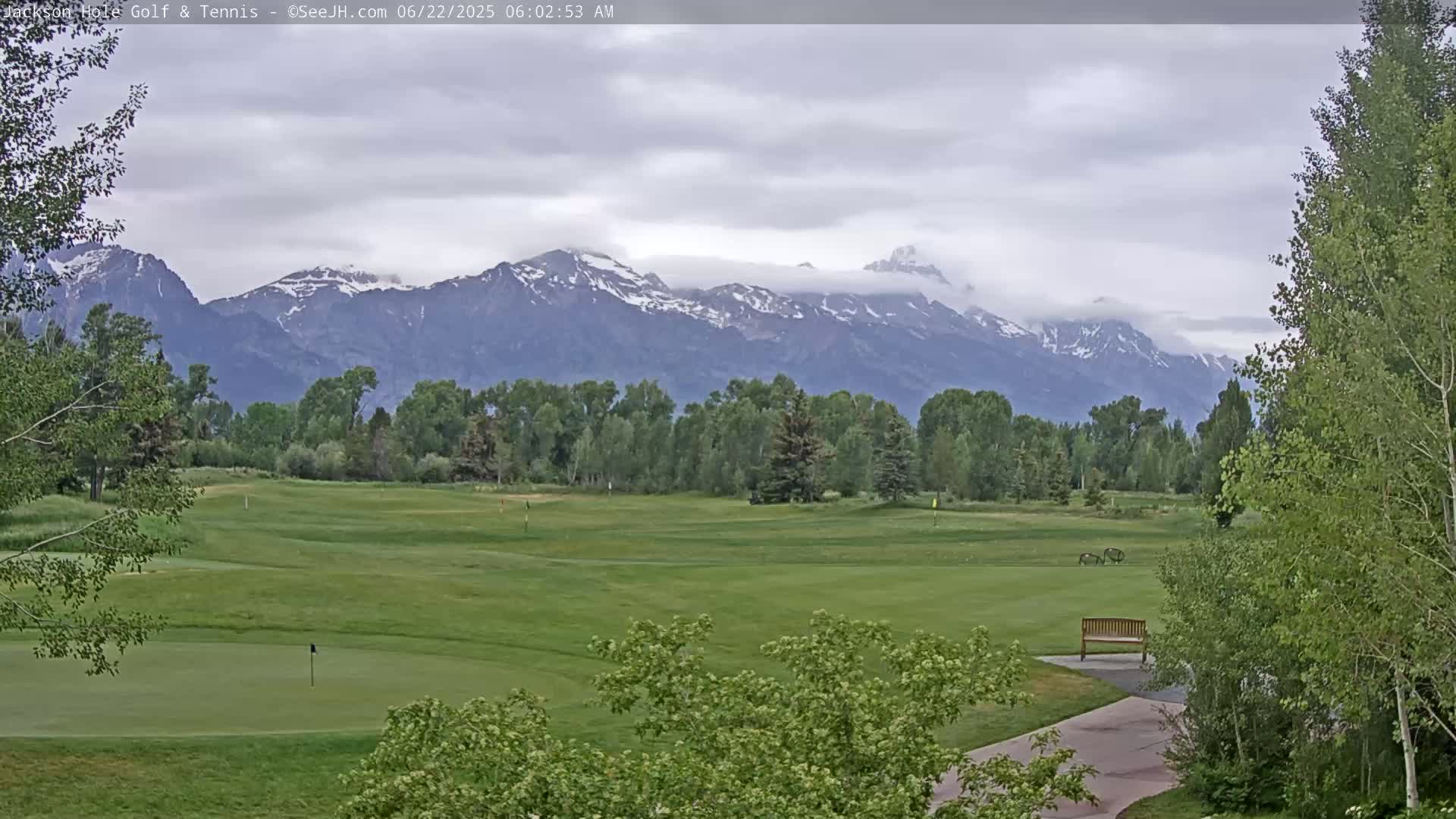 A golf course with a putting green, bordered by green trees, sits in the foreground with a snow-capped mountain range under a cloudy sky in the background.