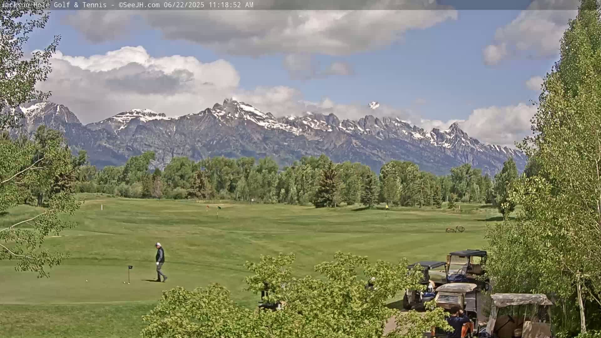 A golfer walks across a green putting surface on a golf course with a partly cloudy sky and snow-capped mountains in the background.