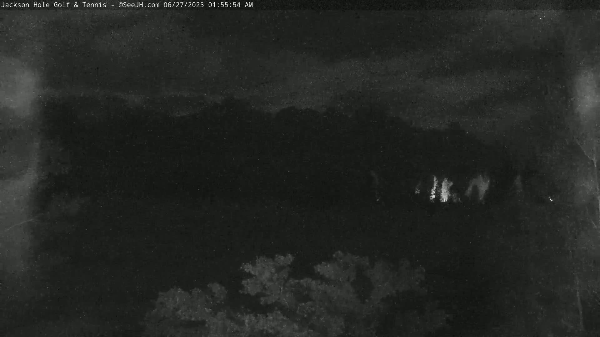 A nighttime, low-light image shows a dark landscape with some faintly illuminated areas in the middle distance and darker vegetation in the foreground.