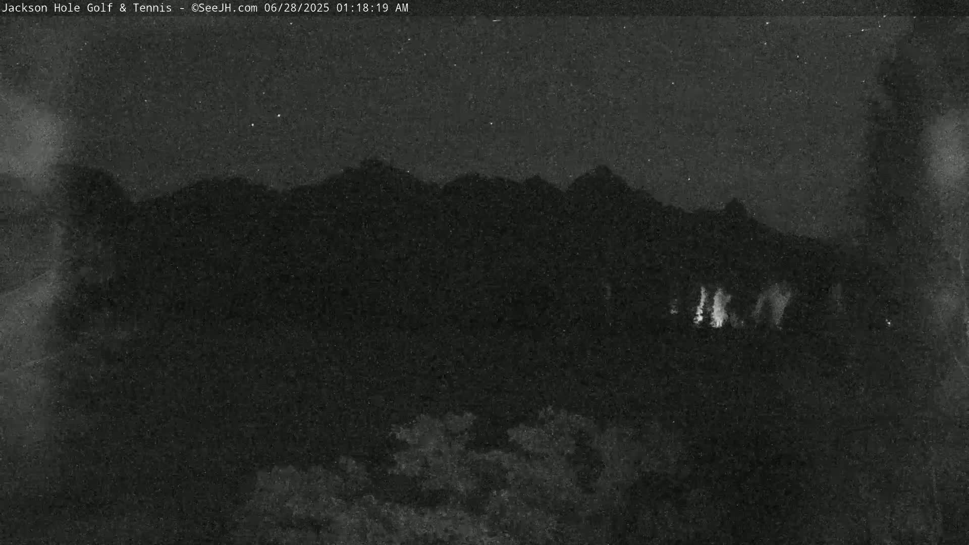 A nighttime view of dark mountains under a clear, starry sky, with faint lights visible in the lower center.