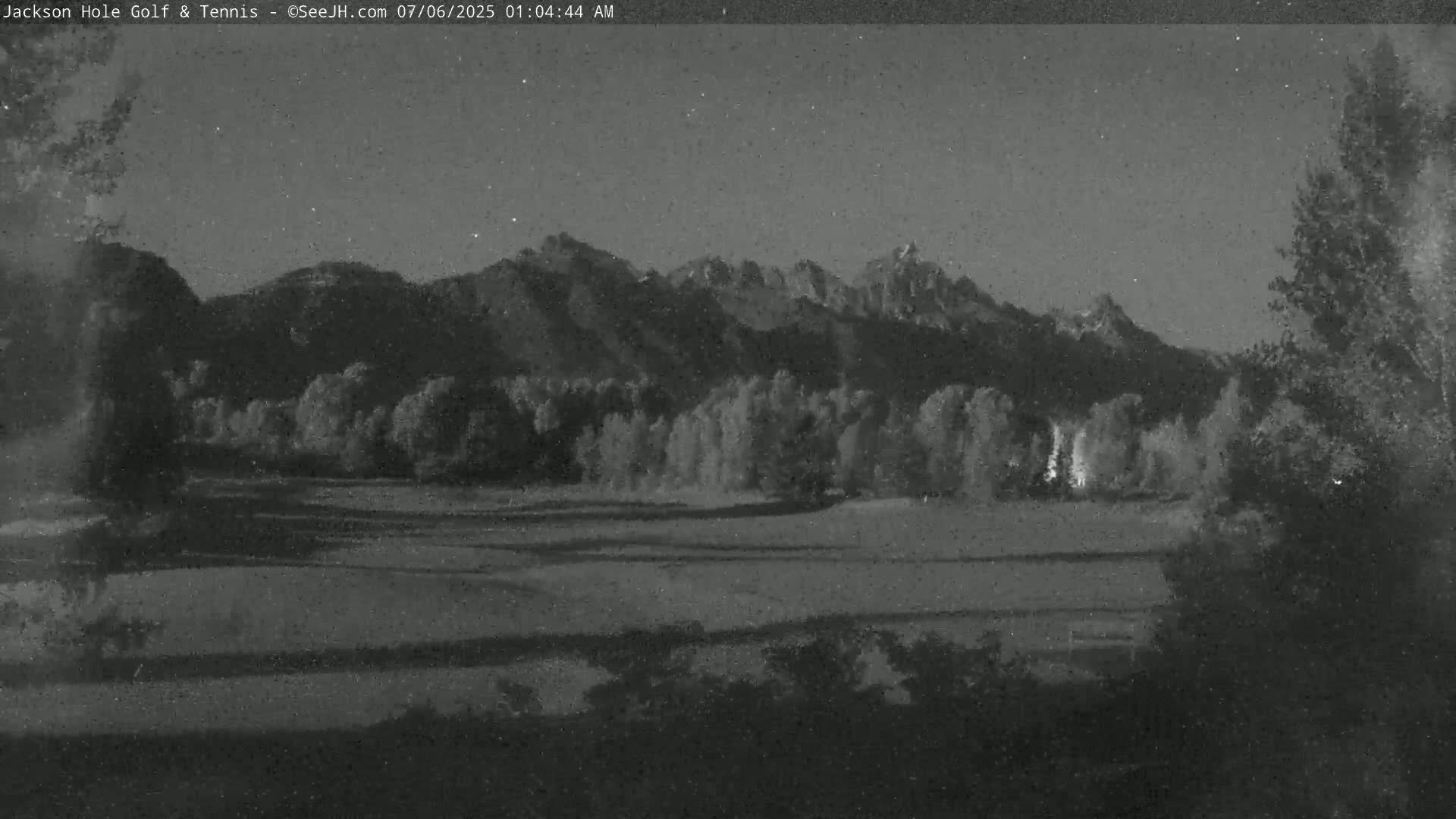 A nighttime view shows a flat field, trees, and a mountain range under a dark, starry sky.