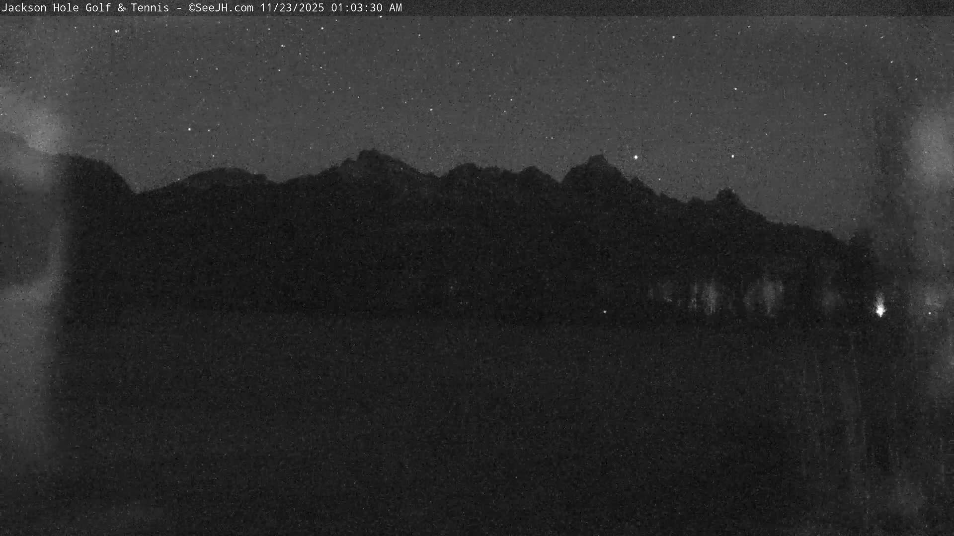 A grainy, dark image shows a mountain range silhouetted against a clear, starry night sky with reflections of distant lights on a body of water below.