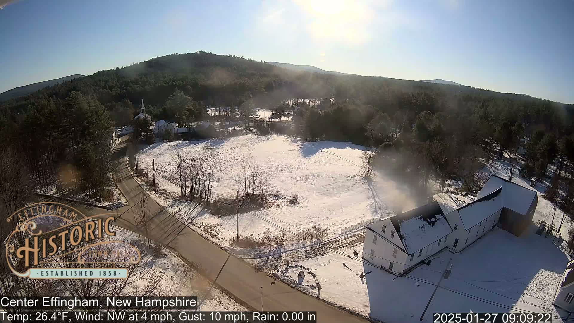 A snow-covered town with a road and buildings is nestled among a wooded hillside under a sunny sky.