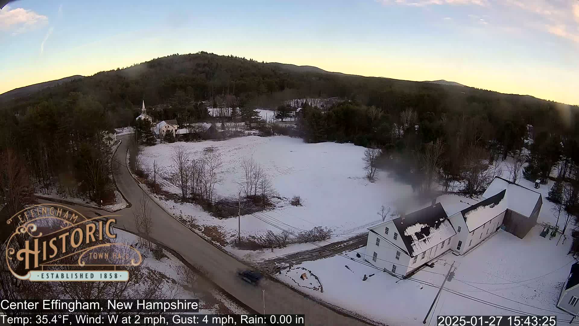 A snow-covered town in a valley, with a road and several buildings visible, under a partly cloudy sky.