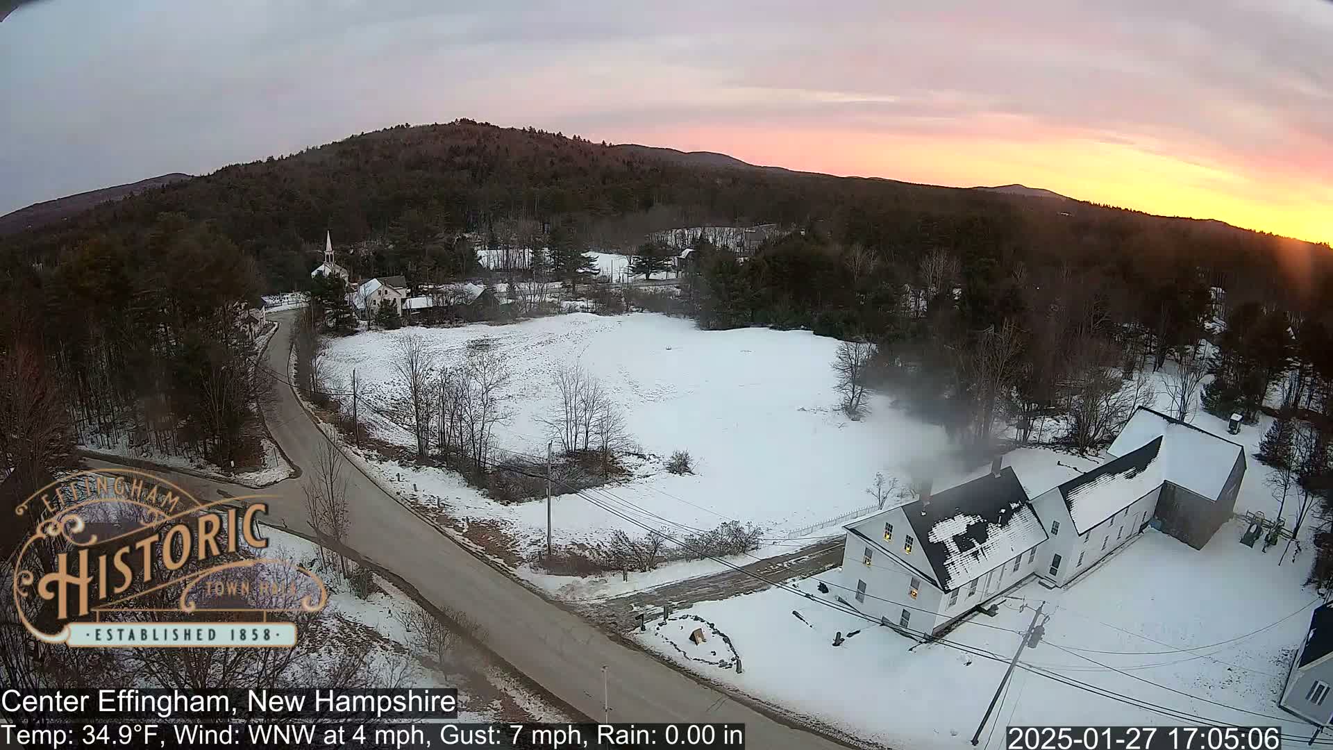 A snow-covered town at sunset, with a road winding through the scene towards hills in the background.
