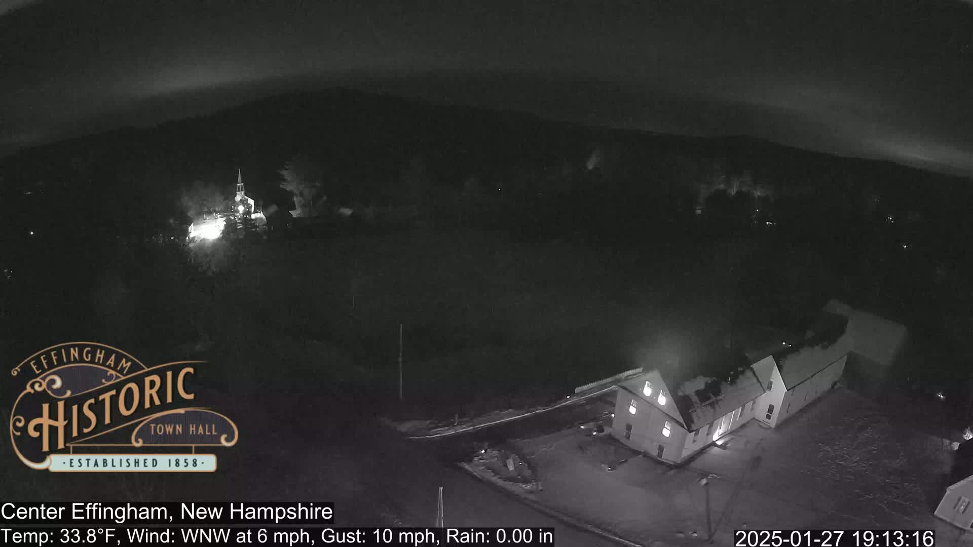 A nighttime, aerial view of a town shows a building illuminated, with a slightly overcast sky and light wind.