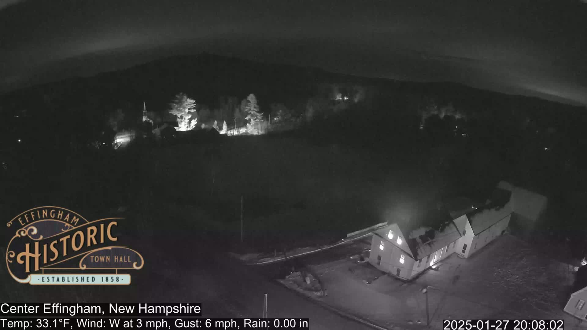 A nighttime, high-angle view of a town with a building illuminated, under a partly cloudy sky with a westerly wind at 3 mph and gusts up to 6 mph.