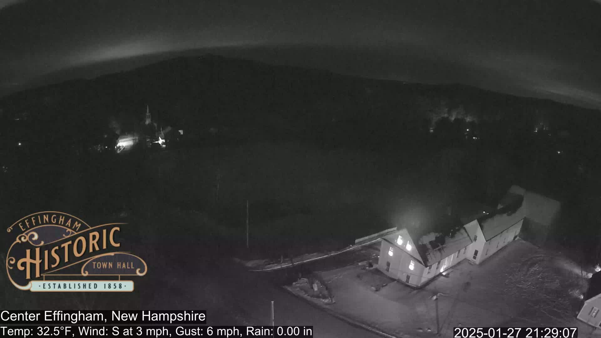 A nighttime, overhead view of a town with some snow on the ground, under a dark sky with a slight glow on the horizon.