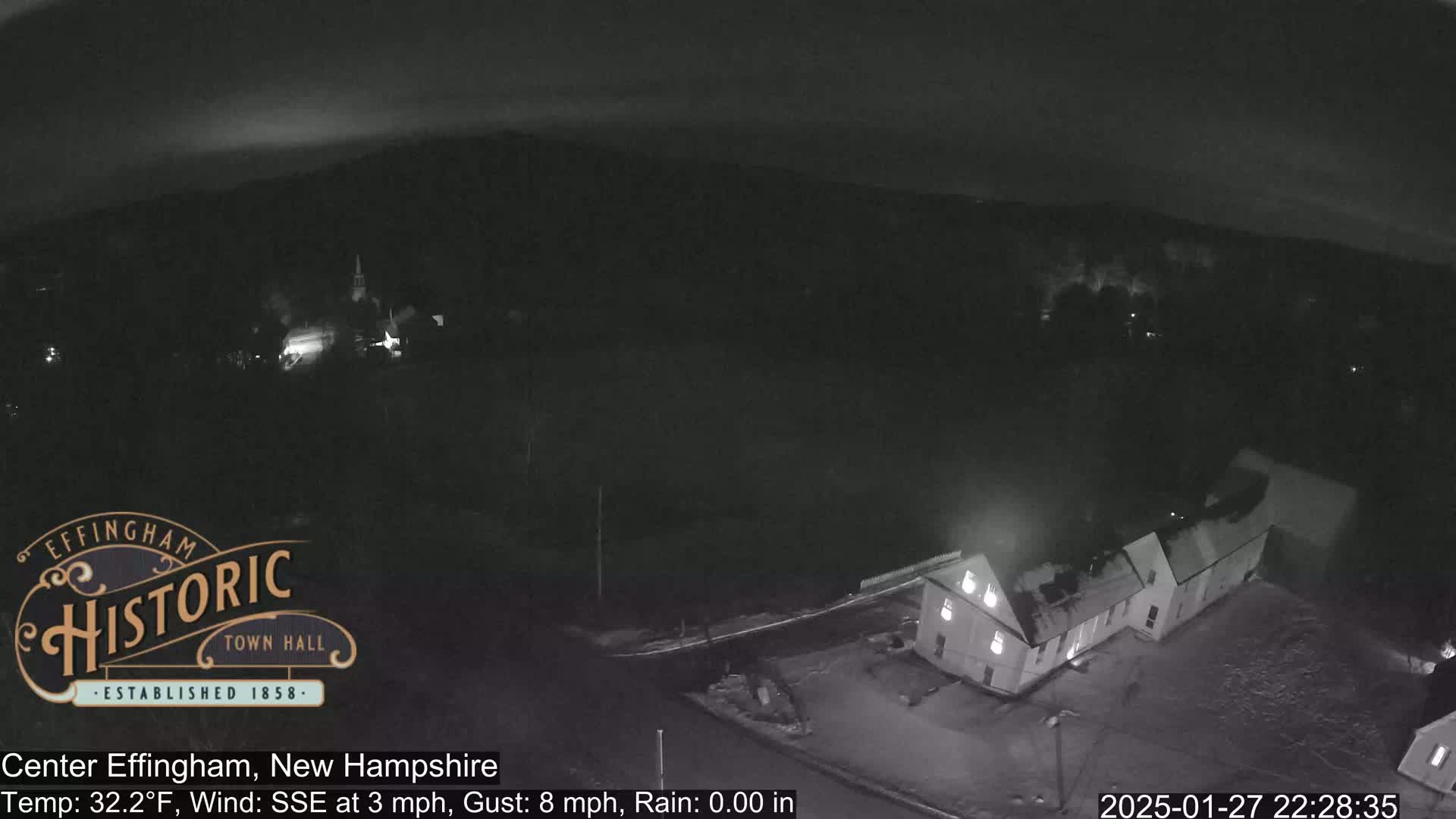 A nighttime, high-angle view of a small town with some buildings and a light dusting of snow on the ground, under a dark, clear sky.