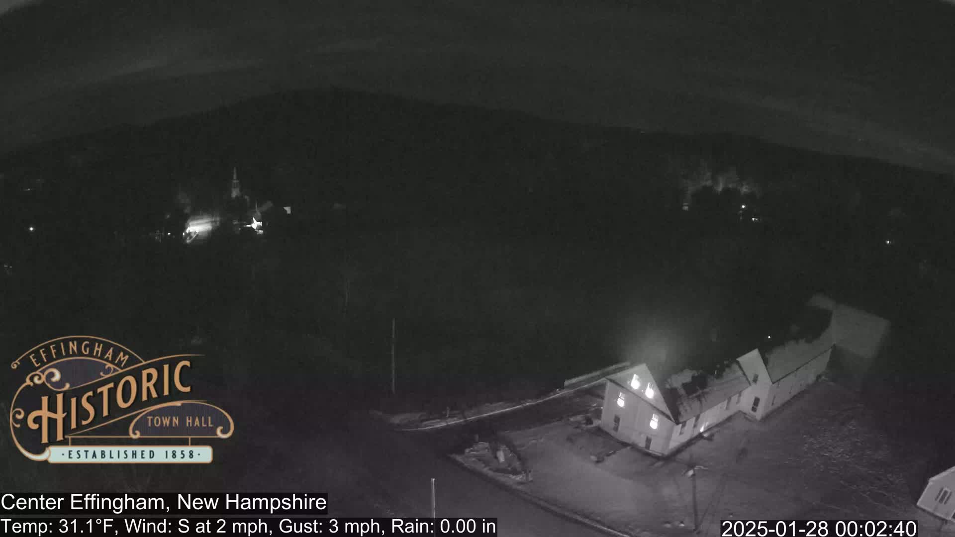 A nighttime, aerial view of a small town under a dark sky with light rain shows a building illuminated in the foreground.