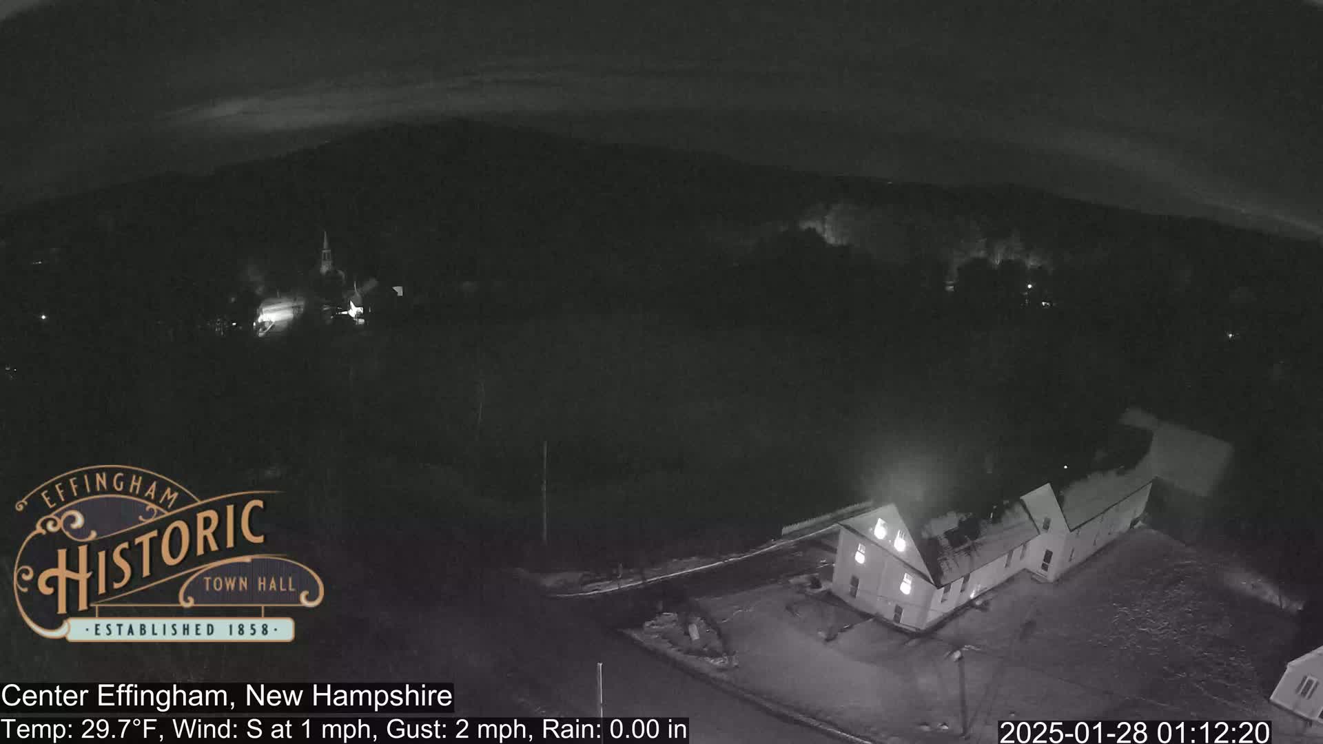 A nighttime, rainy view of a town with a building in the foreground and a church spire in the distance.