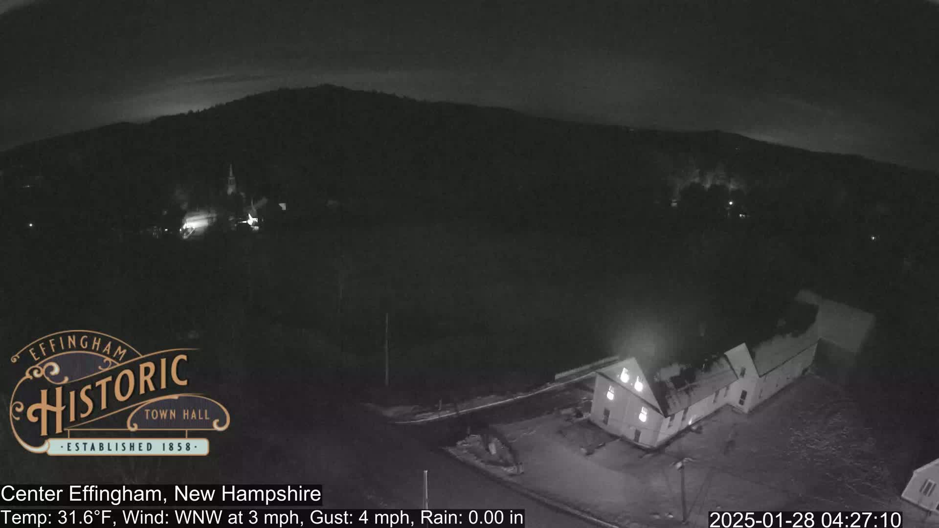 A nighttime, rainy view of a small town with a few buildings, including one with lights on, nestled beneath dark hills.