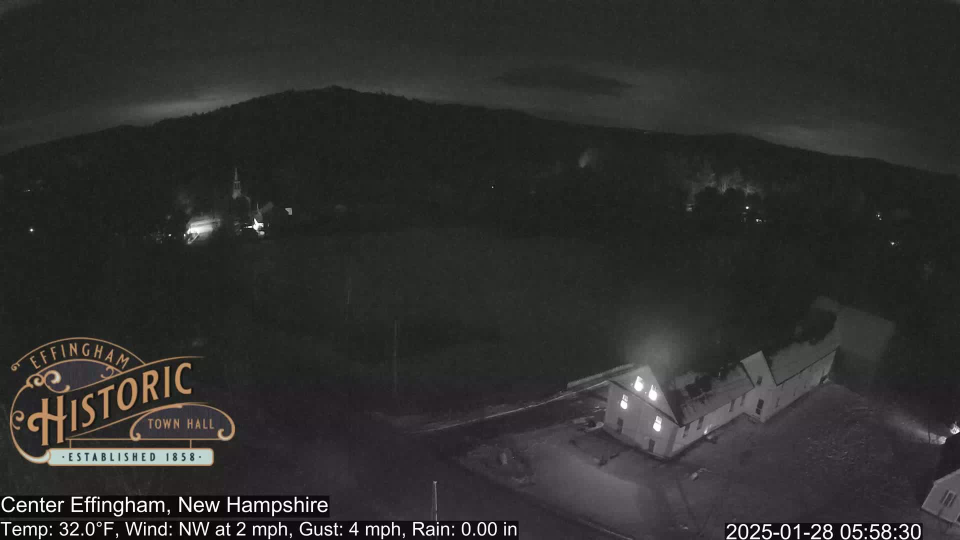 A nighttime, low-light view of a town with a building prominently featured in the foreground under a dark, slightly cloudy sky;  there is a light northwest wind and no rain.